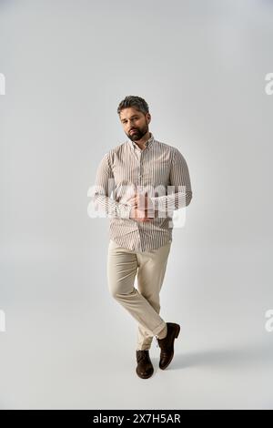 Un homme à la mode avec une barbe portant une chemise rayée et un pantalon kaki pose élégamment sur un fond gris dans un studio. Banque D'Images