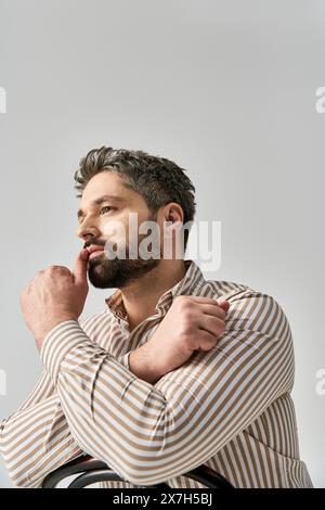 Un homme sophistiqué avec une barbe pose en toute confiance dans une chemise rayée sur un fond de studio gris. Banque D'Images