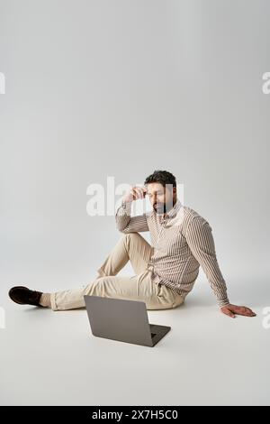 Un homme à la mode avec une barbe est assis sur le sol, portant une tenue élégante, car il travaille intensément sur son ordinateur portable. Banque D'Images