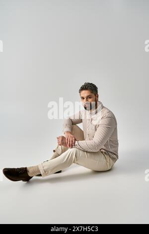 Un homme barbu en tenue élégante assis sur le sol avec ses jambes croisées dans une pose contemplative sur un fond gris de studio. Banque D'Images