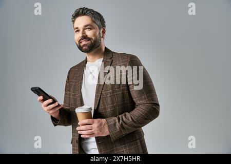 Un homme barbu dans une tenue élégante tient une tasse de café tout en vérifiant son téléphone portable, exsudant sophistication et multitâche. Banque D'Images