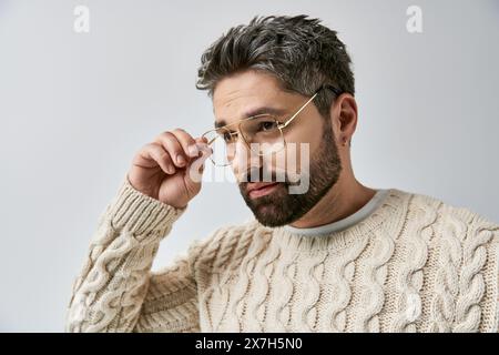 Un homme charismatique avec une barbe pose dans un pull blanc sur un fond gris dans un cadre de studio. Banque D'Images