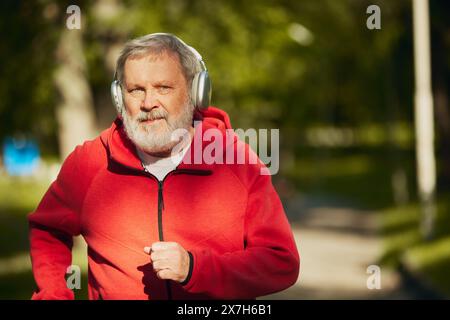 Homme retraité, retraité âgé en sweat à capuche rouge et écouteurs jogging dans le parc tôt le matin ensoleillé. Rester actif Banque D'Images