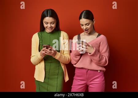 Deux jolies adolescentes brunes se tiennent ensemble, absorbées dans leurs téléphones portables, ignorant leur environnement. Banque D'Images