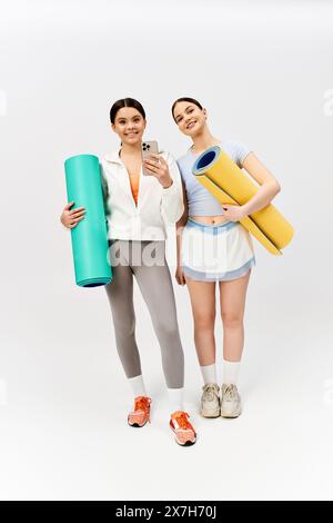 Deux jolies adolescentes, une brune, debout ensemble dans une tenue sportive tenant des tapis de yoga sur fond gris studio. Banque D'Images