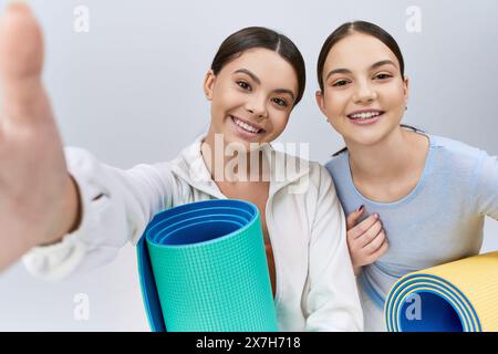 Deux jolies et brunes adolescentes, amies féminines, en tenue sportive debout à côté de l'autre avec des tapis de yoga dans un studio sur un fond gris. Banque D'Images