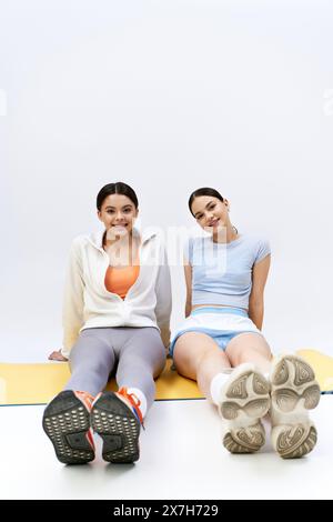 Deux jolies et brunes adolescentes en tenue sportive s'assoient ensemble sur un tapis avec leurs pieds vers le haut dans un cadre de studio. Banque D'Images