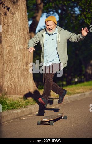 Old School Skater. Homme senior dans des vêtements élégants décontractés profitant d'une matinée active dans le parc, skateboard sur le chemin vide. Banque D'Images