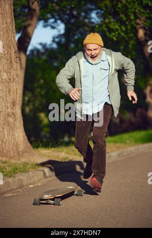 Homme âgé dans des vêtements élégants décontractés skateboard dans le parc du matin le jour ensoleillé, cascades de formation. Activité juvénile Banque D'Images