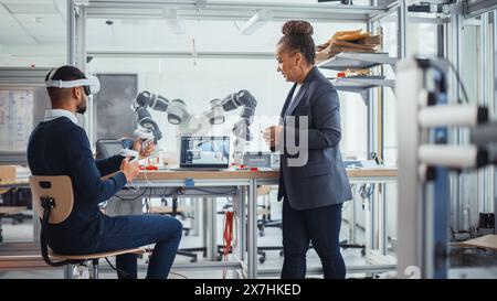 Black Senior Engineer parle à un jeune informaticien masculin qui porte un casque de réalité virtuelle et utilise des contrôleurs pour manipuler à distance la main robotique. Concept de laboratoire de recherche de haute technologie Banque D'Images