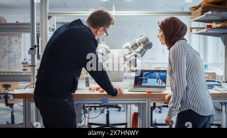 Ingénieur senior expliquant les contrôles à une spécialiste musulmane en informatique féminine et pointant vers la main robotique dans le laboratoire de recherche avec ordinateur portable. Concept innovant de Startup Company. Banque D'Images