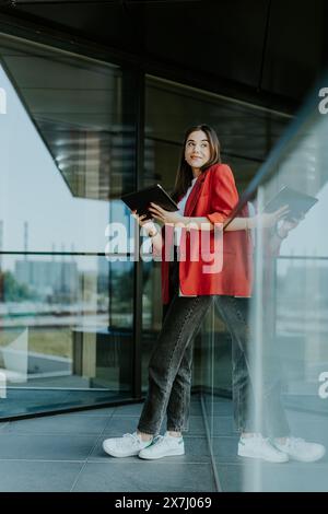 Femme d’affaires joyeuse, vêtue d’un blazer rouge, interagit avec une tablette près d’une grande fenêtre de bureau offrant une vue dégagée sur la ville par une journée ensoleillée Banque D'Images