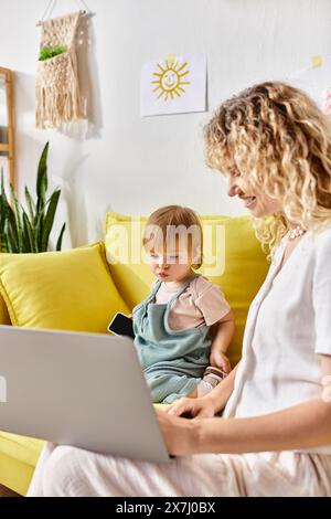 Une mère aux cheveux bouclés assise sur un canapé avec sa fille en bas âge sur ses genoux, partageant un moment d'amour et de tendresse à la maison. Banque D'Images