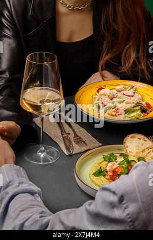 Couple élégant partageant un moment autour d'une salade César gourmande et d'un houmous crémeux aux crevettes et aux légumes verts, accompagnés d'un verre de vin blanc, dans l'élégant roma Banque D'Images