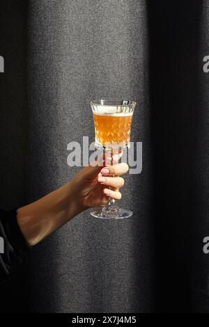 Main féminine tenant élégamment le verre de cristal rempli de whisky aigre, cocktail dans la teinte ambre contrastant avec fond de tissu texturé foncé Banque D'Images