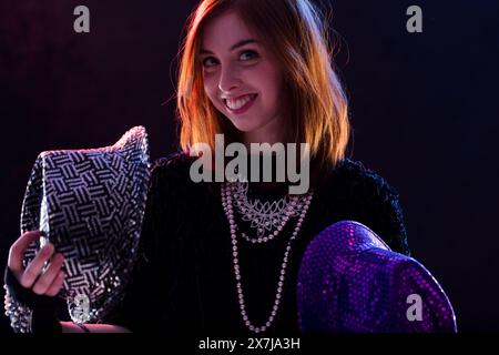 Souriante femme aux cheveux auburn tient un chapeau noir pailleté et un chapeau violet dans un environnement faiblement éclairé. Ses colliers superposés et sa coupe pleine d'assurance Banque D'Images