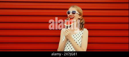 Portrait d'été de jeune femme souriante heureuse avec des fruits juteux, sucette tranche de pastèque portant des lunettes de soleil en forme de coeur avec coiffure girly cool Banque D'Images