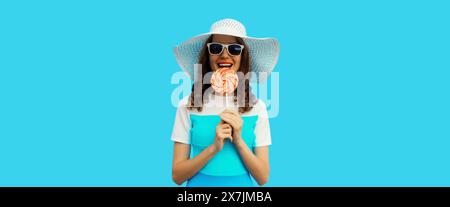 Portrait de jeune femme joyeuse souriante tenant une sucette colorée portant un chapeau d'été blanc, lunettes de soleil sur fond bleu Banque D'Images