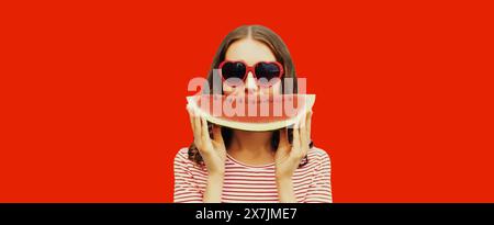 Portrait d'été de jeune femme heureuse avec tranche de pastèque portant des lunettes de soleil en forme de coeur rouge sur fond de studio Banque D'Images