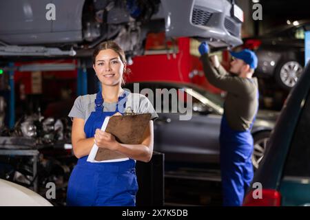 Travailleuse de service automobile tient des registres du travail effectué par les mécaniciens automobiles dans la station-service automobile - prend des notes sur papier Banque D'Images