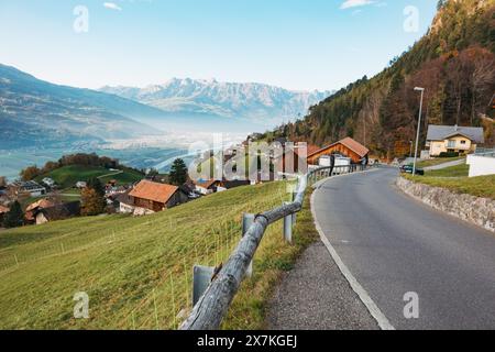 Une route sinueuse mène à travers un village pittoresque du Liechtenstein niché dans les contreforts des Alpes, avec une vue sur une vallée et des montagnes lointaines Banque D'Images
