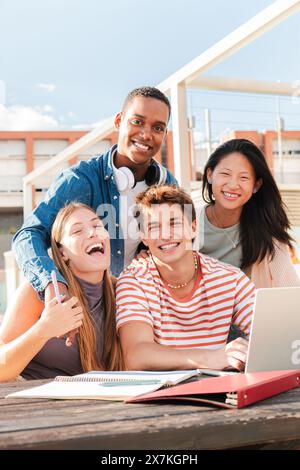 Vertical. Groupe diversifié d'étudiants masculins et féminins regardant la caméra tout en étudiant en collaboration à l'aide d'un ordinateur portable sur le campus de l'Université. Adolescents multiraciaux souriant et faisant les devoirs. Photo de haute qualité Banque D'Images