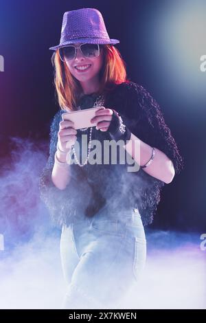 Sous les lumières de la scène, une jeune femme aux cheveux roux, chapeau à paillettes et lunettes de soleil utilise son téléphone, paraissant pleinement engagée et heureuse, habillée dans un bla chic Banque D'Images