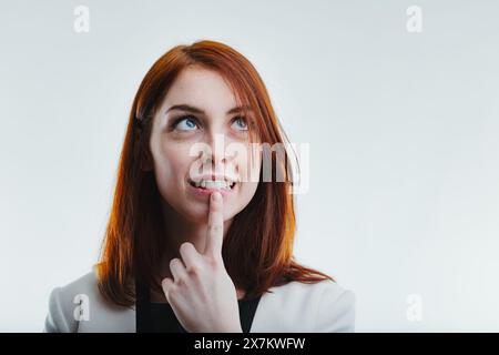 Rousse réfléchie avec une expression curieuse, touchant doucement son menton, vêtue d'un blazer blanc sophistiqué Banque D'Images