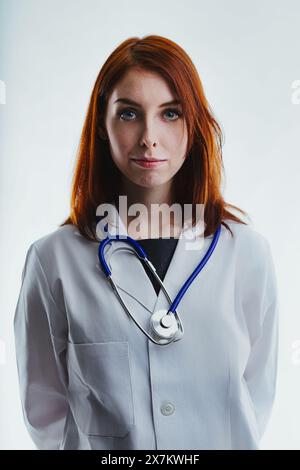 médecin aux cheveux roux portant une blouse de laboratoire blanche et un stéthoscope autour du cou regarde directement la caméra. Son expression sérieuse le souligne Banque D'Images