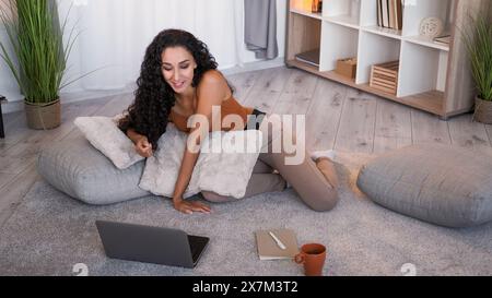 Appel vidéo. Chat en ligne. Communication Internet. Femme souriante détendue parlant sur ordinateur portable sur le sol à l'intérieur du salon à la maison avec espace libre. Banque D'Images