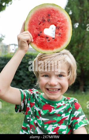 joyeux garçon mange une pastèque, découpant un cœur dans un morceau rond de pastèque Banque D'Images