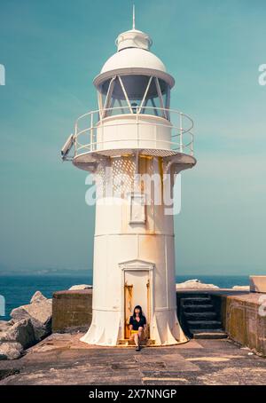 Femme en noir et jaune assise au pied d'un phare blanc avec un fond de ciel bleu sarcelle, Brixham Lighthouse, Devon, Royaume-Uni Banque D'Images