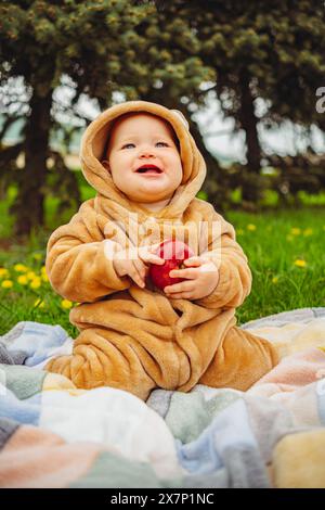 Mignon bébé fille en costume en peluche avec pomme rouge dans les mains assis sur une couverture dans l'herbe verte Banque D'Images