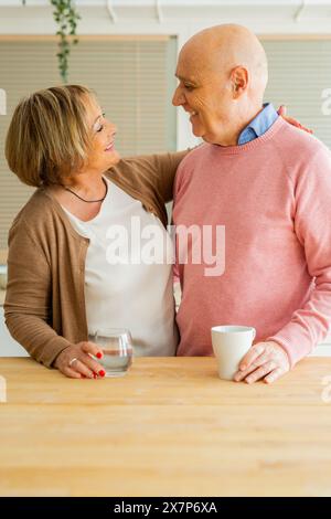 Portrait d'un couple d'aînés souriant, se regardant, debout ensemble dans une cuisine avec un verre d'eau et une tasse blanche. Banque D'Images