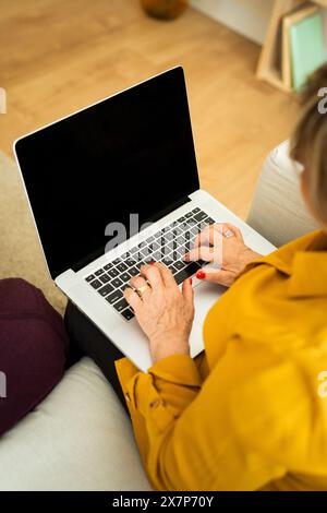 Gros plan d'une femme âgée tapant sur un ordinateur portable. Mains de la femme senior sur le clavier dans un cadre confortable à la maison. Banque D'Images