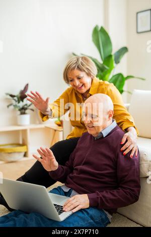 Couple senior ayant un appel vidéo sur un ordinateur portable, souriant et agitant à l'écran. Couple senior dans un salon, se connectant avec des proches en ligne. Banque D'Images