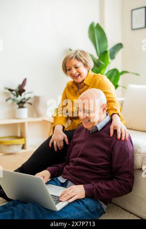 Couple senior ayant un appel vidéo sur un ordinateur portable, souriant à l'écran. Couple senior dans un salon, se connectant avec des proches en ligne. Banque D'Images