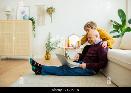 Couple senior ayant un appel vidéo sur un ordinateur portable, souriant et agitant à l'écran. Couple senior dans un salon, se connectant avec des proches en ligne. Banque D'Images