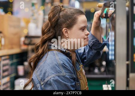 Vue de côté portrait d'une jeune femme atteinte du syndrome de Downs travaillant dans la boulangerie et regardant la pâtisserie dans le four profitant de la vie indépendante Banque D'Images