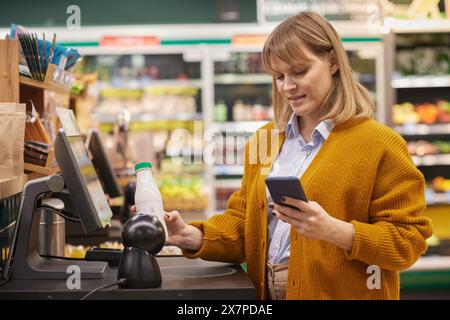 Portrait de taille vers le haut de femme adulte blonde scannant les produits laitiers tout en utilisant le service de caisse libre-service et tenant le téléphone dans le supermarché Banque D'Images