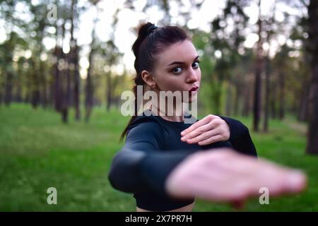 Jeune femme faisant la formation d'arts martiaux dans le haut noir sportif tenant les mains dans la pose de combat sur fond de forêt. Banque D'Images