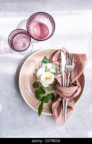 Une configuration de salle à manger surélevée avec une assiette rose pâle avec une serviette en terre cuite soigneusement pliée, des couverts et une rose blanche Banque D'Images