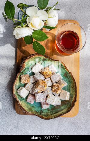 Un mélange de délices turcs saupoudrés sur une assiette en céramique avec un verre de thé noir, accompagné d'un vase de roses blanches, le tout présenté sur une planche de bois Banque D'Images