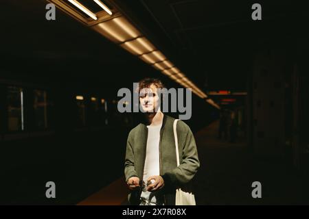 Un jeune homme se tient debout sur une plate-forme de métro faiblement éclairée, baigné dans un éclat dramatique de lumière frappant son visage, serrant un sac dans une pensée décontractée Banque D'Images
