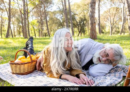 Un joyeux couple de personnes âgées partage un rire tout en étant allongé sur une couverture de pique-nique dans un parc ensoleillé, entouré de grands arbres Banque D'Images