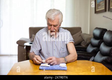 Un homme caucasien âgé se concentrant tout en vérifiant son taux de sucre dans le sang avec un glucomètre à une table en bois dans un salon bien éclairé, highlighti Banque D'Images