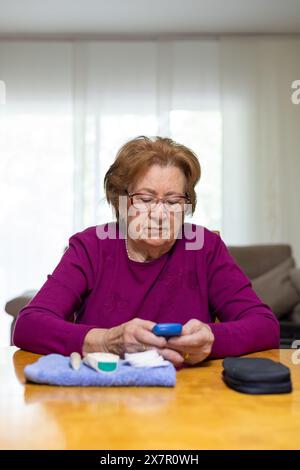Une femme âgée se concentre sur l'utilisation d'un glucomètre pour vérifier son taux de sucre dans le sang à la maison, assise à une table en bois avec des fournitures médicales à proximité, Hig Banque D'Images