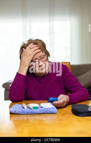 Une femme âgée est assise à une table, visiblement en détresse, alors qu'elle vérifie son taux de sucre dans le sang avec un glucomètre, indiquant un épisode hypoglycémique Banque D'Images