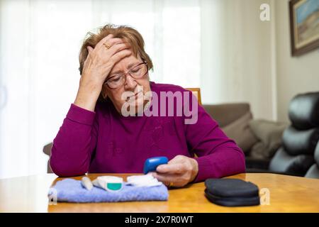 Une femme âgée dans un pull violet semble inquiète alors qu'elle vérifie son taux de glucose dans le sang, indiquant des symptômes d'hypoglycémie Banque D'Images