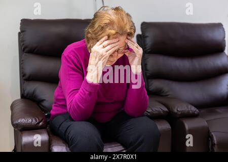 Une femme âgée est assise en détresse sur un canapé, tenant sa tête dans l'inconfort, illustrant les symptômes de l'hypoglycémie Banque D'Images